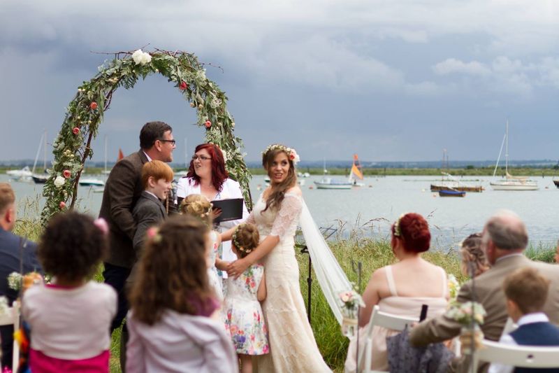 Roxanne leading a wedding under a flower arch beside the water at West Mersea